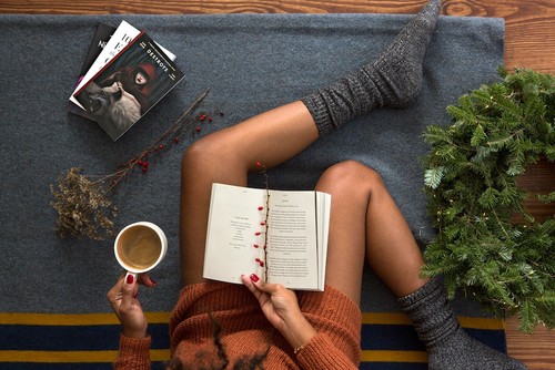 Image of a woman's lap; she's relaxing on cushions with a hot beverage while reading a book